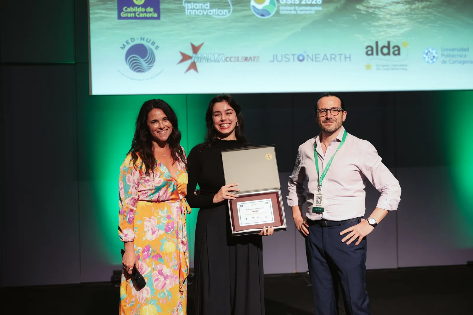 Award winners hold a framed certificate and trophy alongside the host on stage in front of the GSIS 2026 and partner logo wall at the Solutions Showcase, Gran Canaria